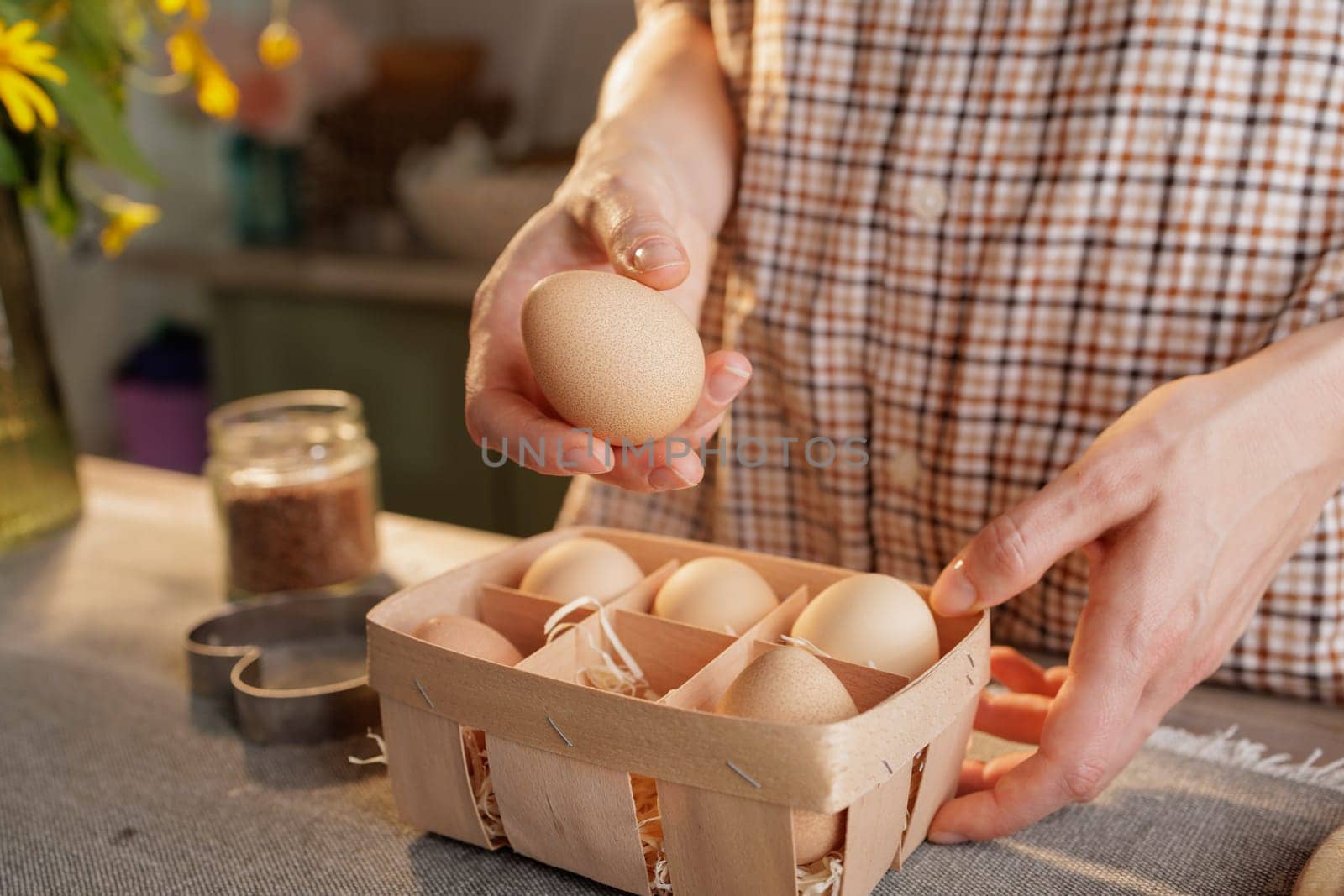 Close-up of woman's hand holding egg over basket with several eggs, kitchen background. Fresh organic eggs concept