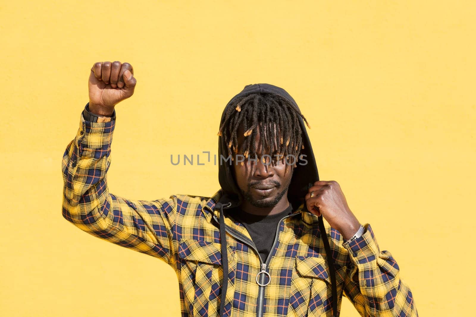attractive young black man with raised fist and hood on an intense yellow background, concept of human rights struggle and lifestyle, copy space for text