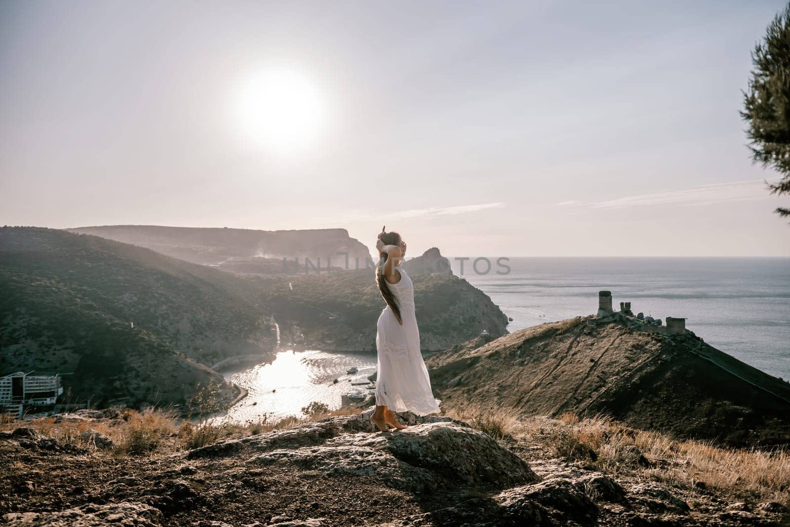 woman stands on a rocky hill overlooking a body of water. She is wearing a white dress and she is enjoying the view