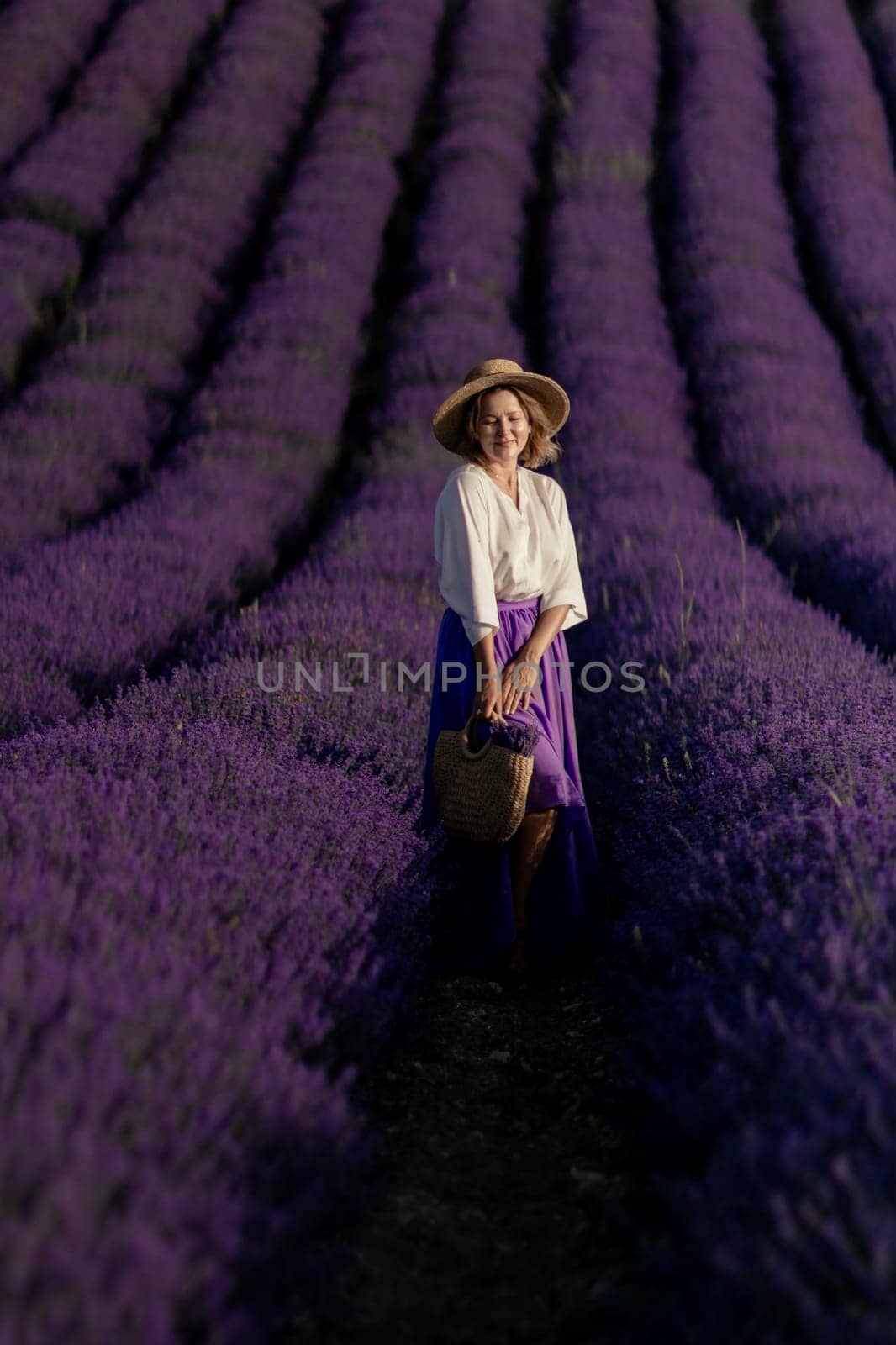 A woman is standing in a field of lavender flowers. She is wearing a straw hat and a blue skirt