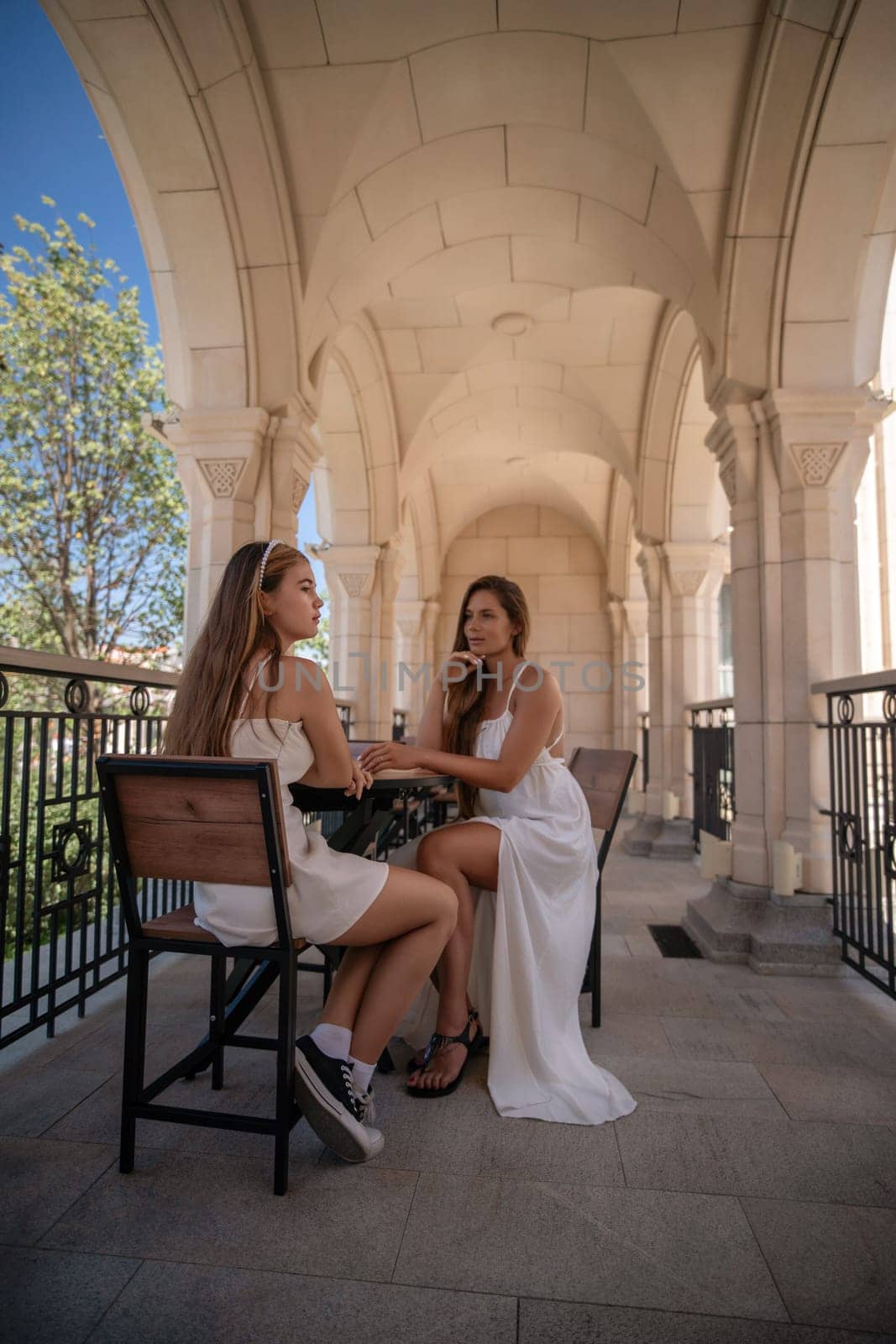 Two women in white dresses sit at a table on a balcony with stone arches, chatting and enjoying the view.