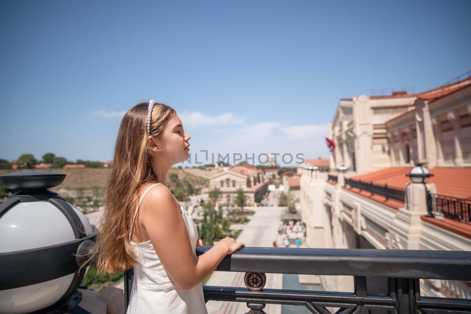 A woman in a white dress stands on a balcony, looking out at a cityscape under a clear blue sky.