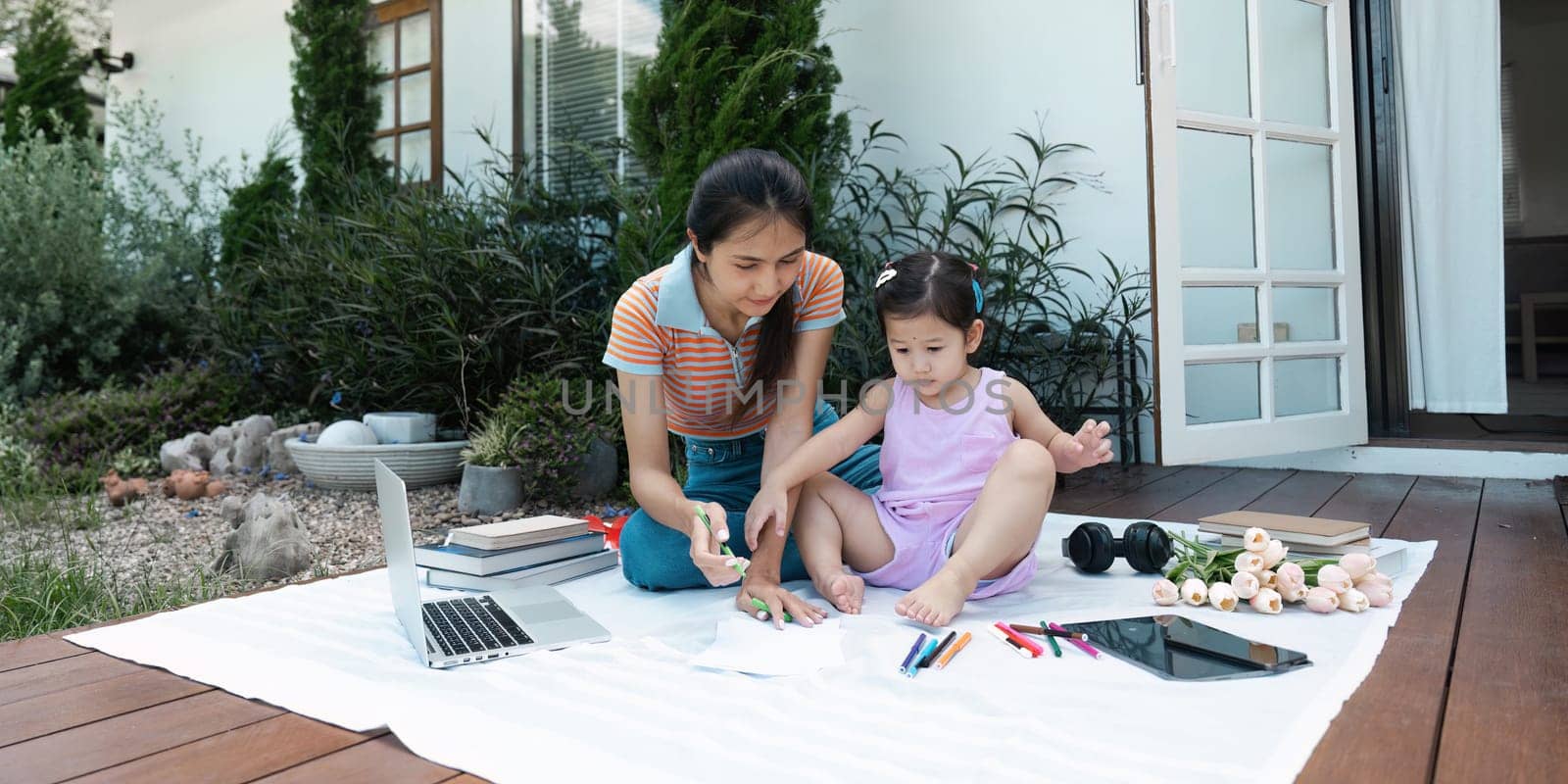 A mother and daughter spending quality time together outdoors, surrounded by art supplies, books, and a laptop on a sunny day.