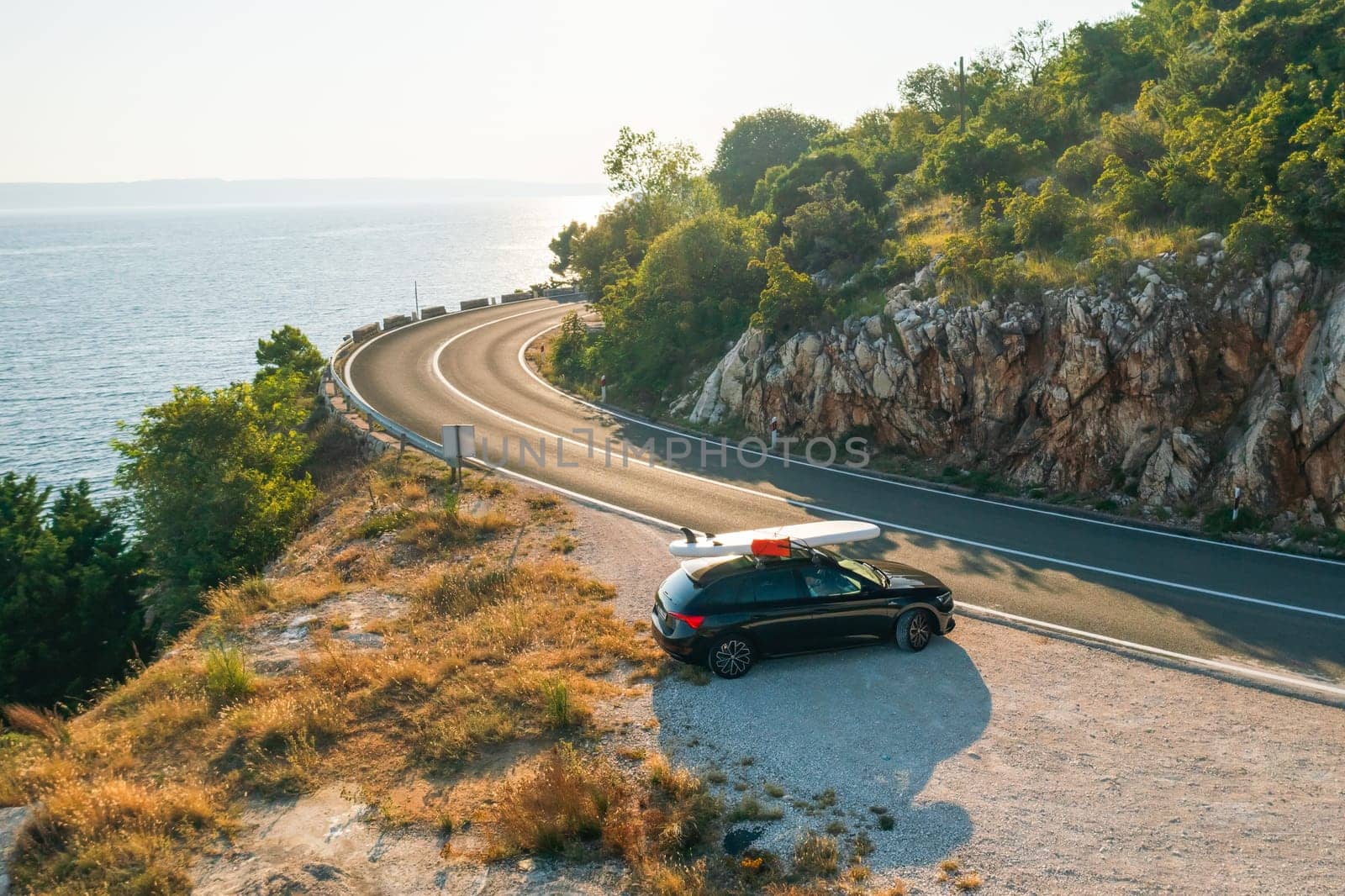 Black car with a paddleboard secured to its roof, parked at a scenic viewpoint on a high cliff