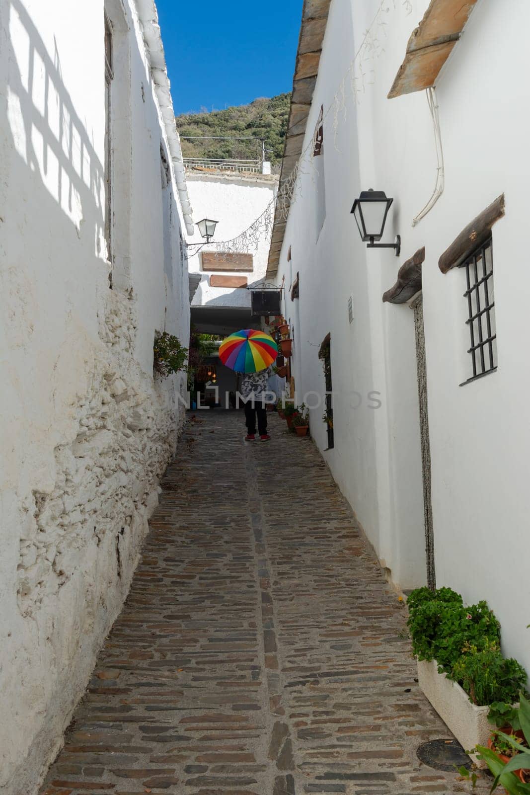 Narrow cobblestone alley lined with white buildings, person holding a colorful umbrella in a sunny scene, Alpujarras, granada, andalucia, espa a,