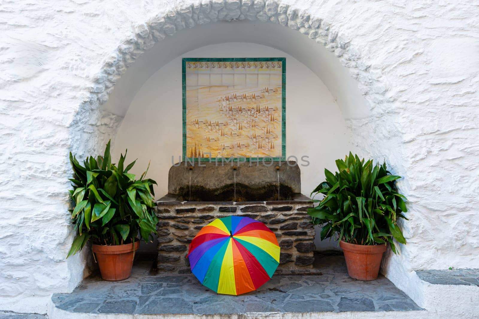 A colorful rainbow umbrella placed in front of an arch with white walls, potted plants, and hanging artwork, Alpujarras, granada, andalucia, espa a,