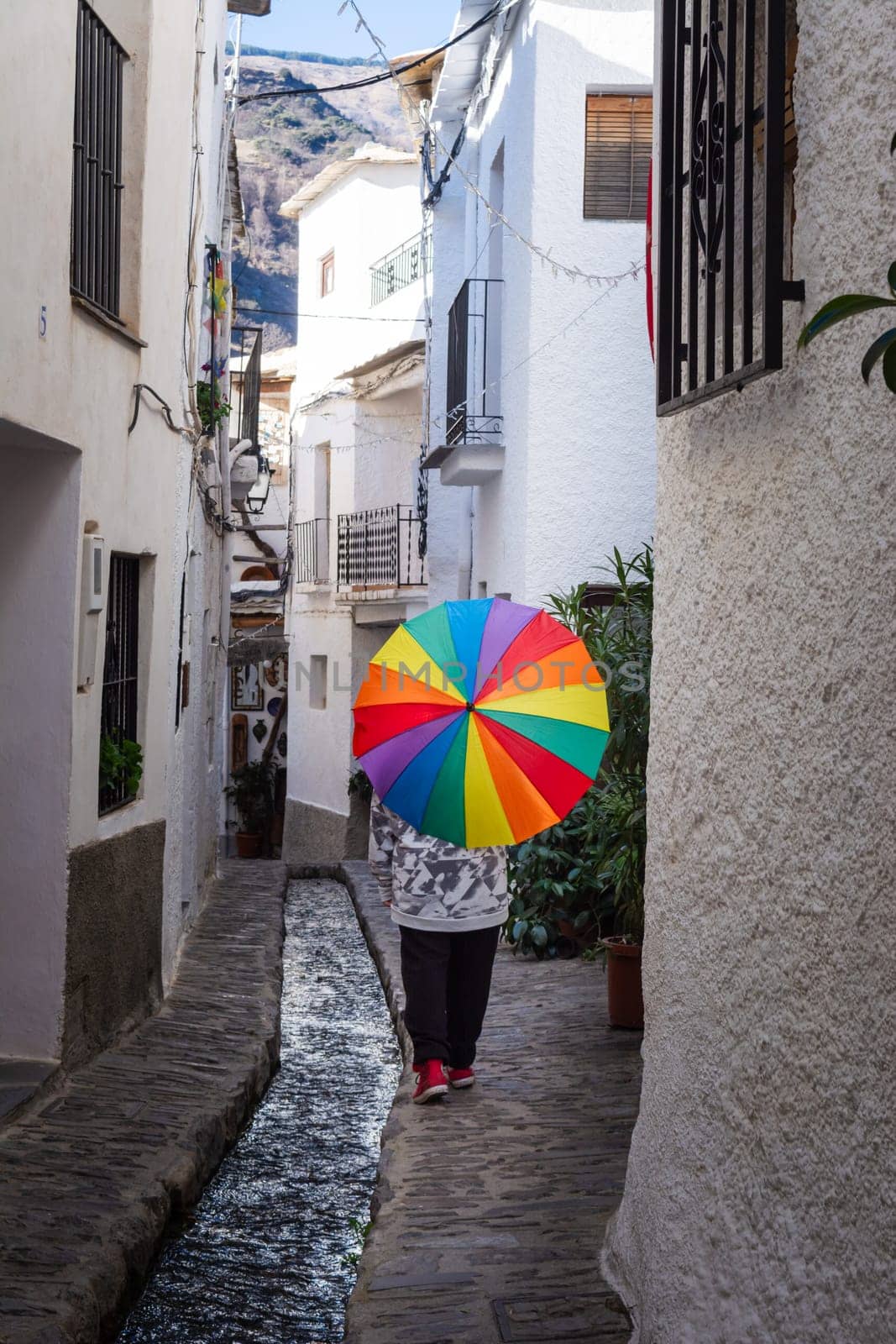 Person holding a colorful umbrella walking in a narrow alley with a water stream and white buildings, Alpujarras, granada, andalucia, espa a,