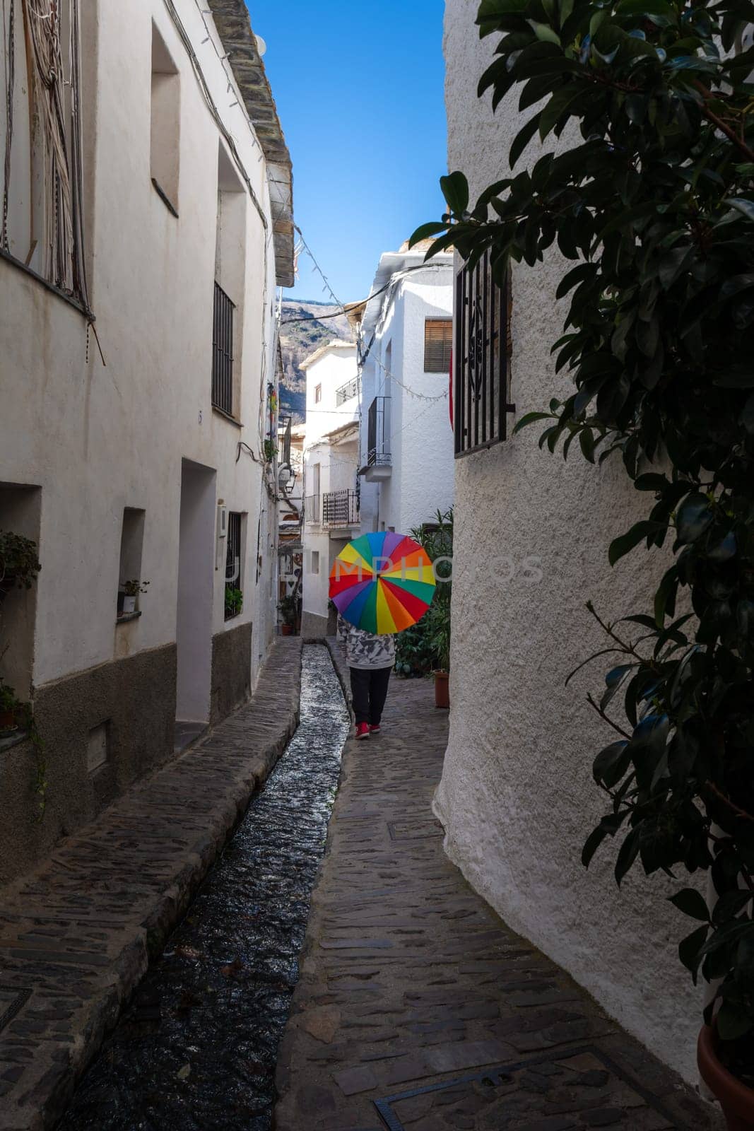 A person with a colorful umbrella walking through a narrow alley with a stream beside white buildings, Alpujarras, granada, andalucia, espa a,