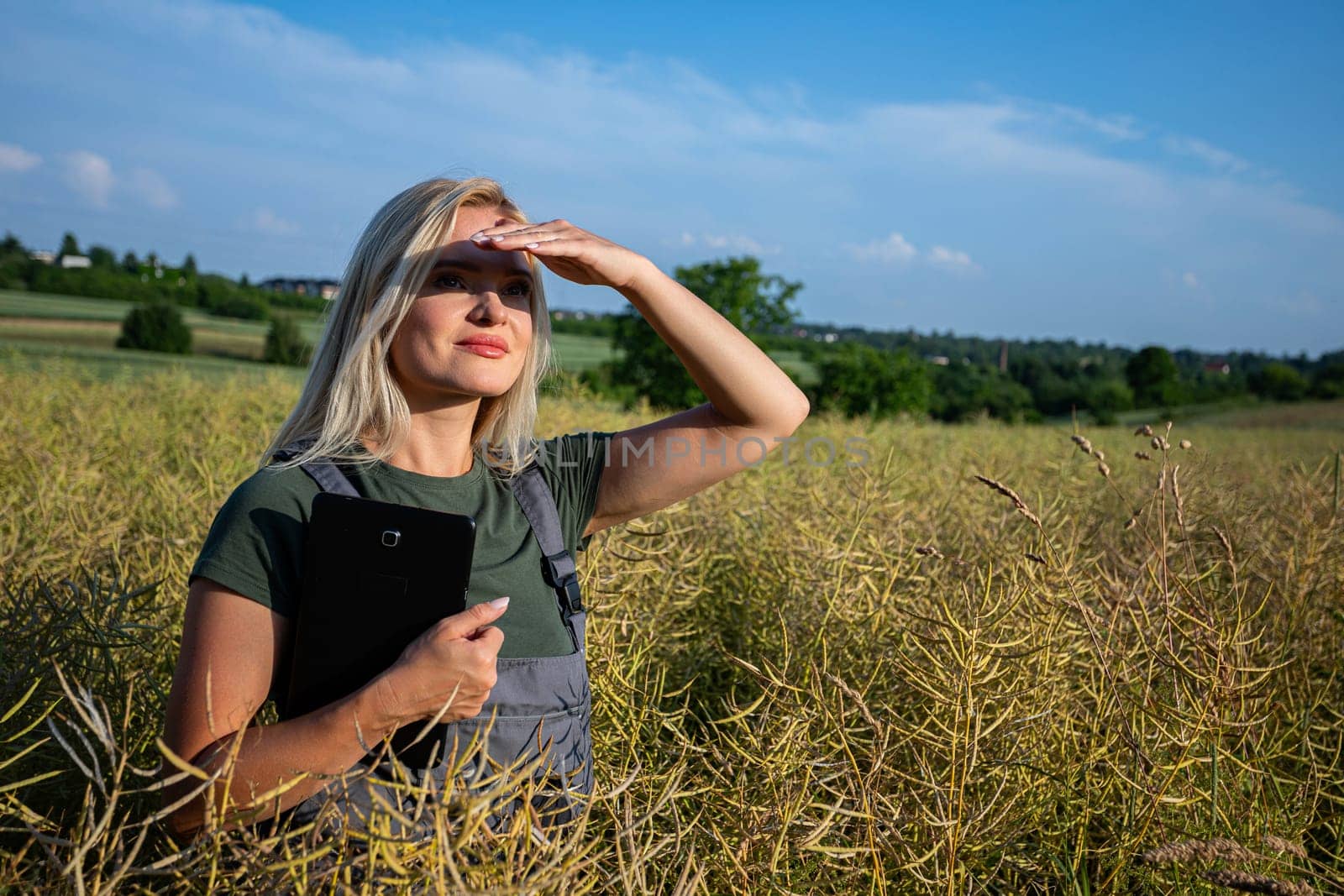In the middle of the rapeseed field, a woman farmer stands, holding a tablet close to her body as she checks the quality of the crop. With one hand on the tablet, she gazes into the distance, her eyes fixed on the blue sky. Experienced and dedicated, she takes pride in caring for her crops in the vast field. She has blonde hair and she smiles with satisfaction, though the sun's rays also glare down upon her, causing her to squint slightly and shield her eyes with her hand. he fertile soil has proven productive once again, yielding yellow rapeseed ready for harvest. With her tablet in hand, the woman farmer embraces the bountiful and lavish harvest. Clad in working clothes, her tanned skin radiates contentment as she stands amidst the fields. Under the blue sky adorned with fluffy white clouds, her spirit merges with the warmth of the sun, celebrating the culmination of her dedication and the richness of the land's yield