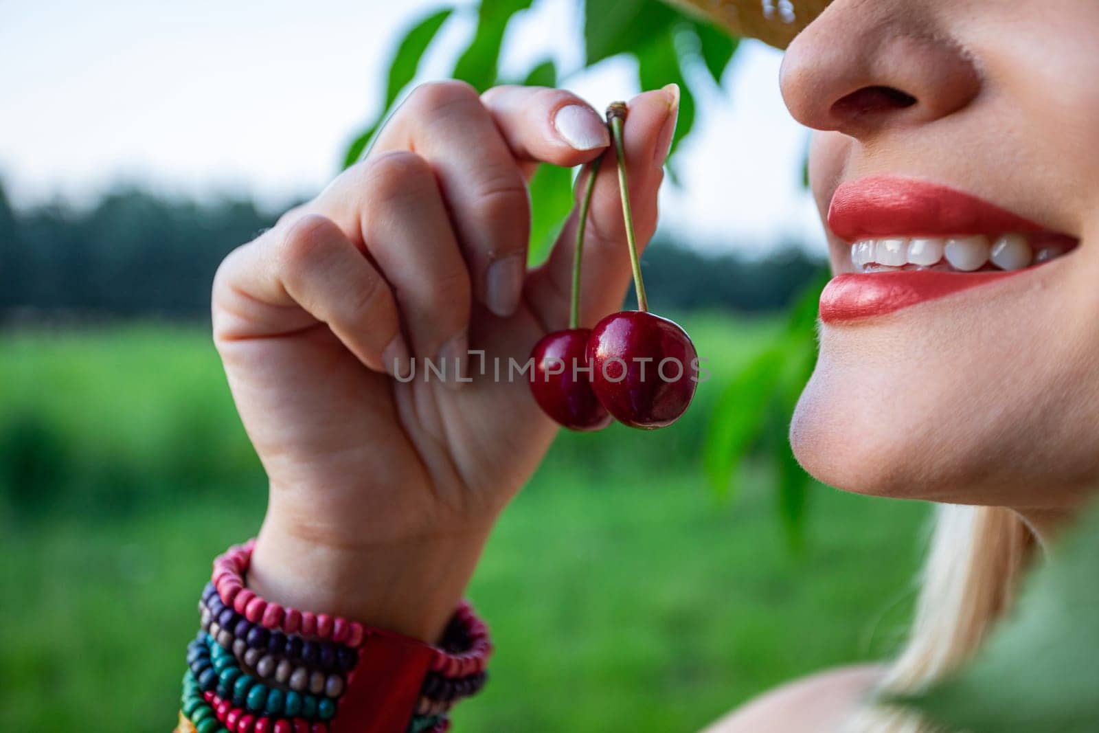 The close-up image captures the essence of a woman's face and hands, revealing her painted, full lips, hands are holding red cherries. Her gaze is directed towards the sky, framed by a backdrop of a cherry tree branch and a blurred forest in the distance. Imperfections adorn her skin, adding a raw authenticity to her presence, while her eyes are barely visible. She has smile on her face showing white teeth. Against her wrist, a colorful bracelet crafted from beads adds a playful touch to her demeanor. The cherries in her grasp exude ripeness, promising a burst of sweetness with every bite. Amidst the tranquil surroundings of the orchard, she stands as a symbol of connection to nature, her contemplative gaze hinting at a moment of quiet reflection amidst the beauty of the natural world
