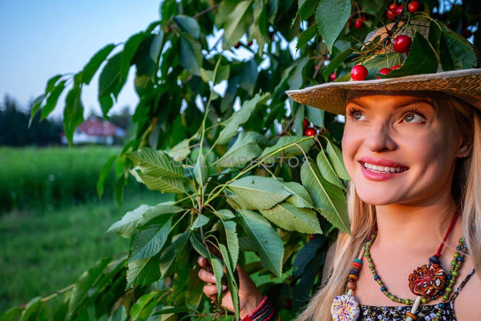 In the serene ambiance of a countryside landscape, a vision of beauty emerges. A radiant woman, her blonde hair framing her face, gazes upwards towards the heavens and the setting sun. The golden rays of sunlight gracefully illuminate her features, accentuating the emerald green of her eyes and the warmth of her smile, revealing her pearly white teeth. Clad in light summer attire, she exudes an air of casual elegance, her ensemble complemented by a colorful beaded necklace adorning her neck and a straw hat atop her head. One hand delicately cradles a branch of the cherry tree, while the other wrist is adorned with a vibrant bracelet. Perched atop her straw hat, a branch of the cherry tree laden with ripe fruits adds a whimsical touch to the scene. Behind her, the cherry tree stands tall, its branches reaching towards the azure sky, while the rural landscape stretches out beyond, a picturesque backdrop to the tranquil moment captured in the photograph. In this idyllic setting, amidst the beauty of nature, the woman finds serenity and joy, a harmonious blend of elegance and rustic charm in the simplicity of the countryside.