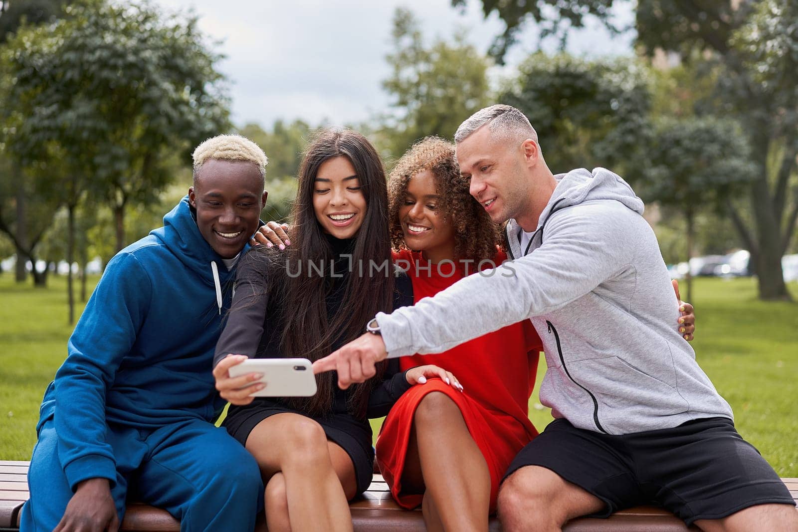 Young people of different nationalities taking selfie using smartphone sitting on bench in park in summer. Happy friends in sportswear spending leisure time together.