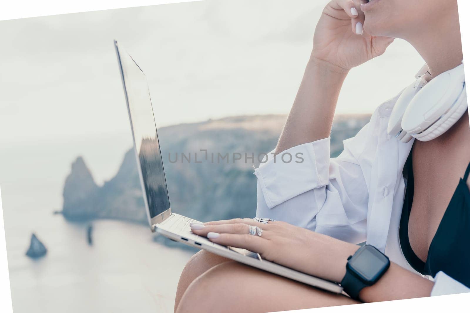 Digital nomad, woman in the hat, a business woman with a laptop sits on the rocks by the sea during sunset, makes a business transaction online from a distance. Freelance, remote work on vacation.