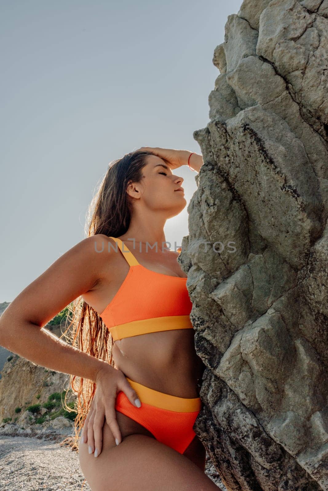 A woman in a bikini is posing in front of a large rock. The rock is grey and has a rough texture. The woman is wearing an orange bikini top and a yellow bikini bottom. She is standing on the beach