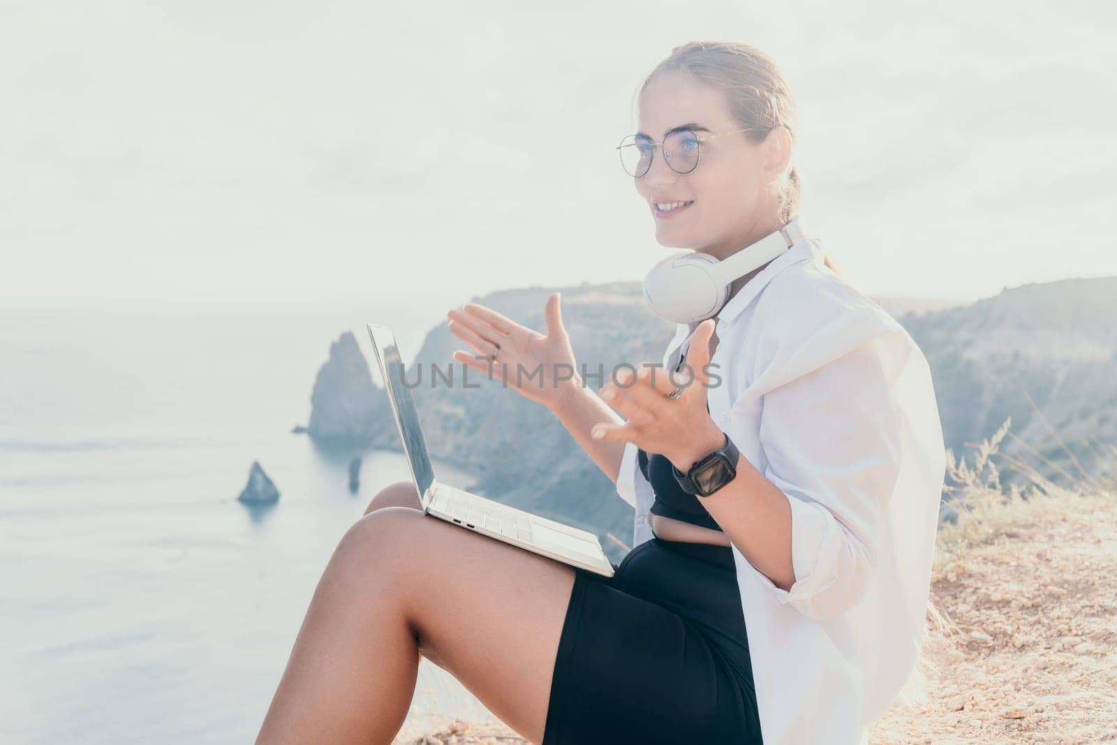 Digital nomad, woman in the hat, a business woman with a laptop sits on the rocks by the sea during sunset, makes a business transaction online from a distance. Freelance, remote work on vacation.