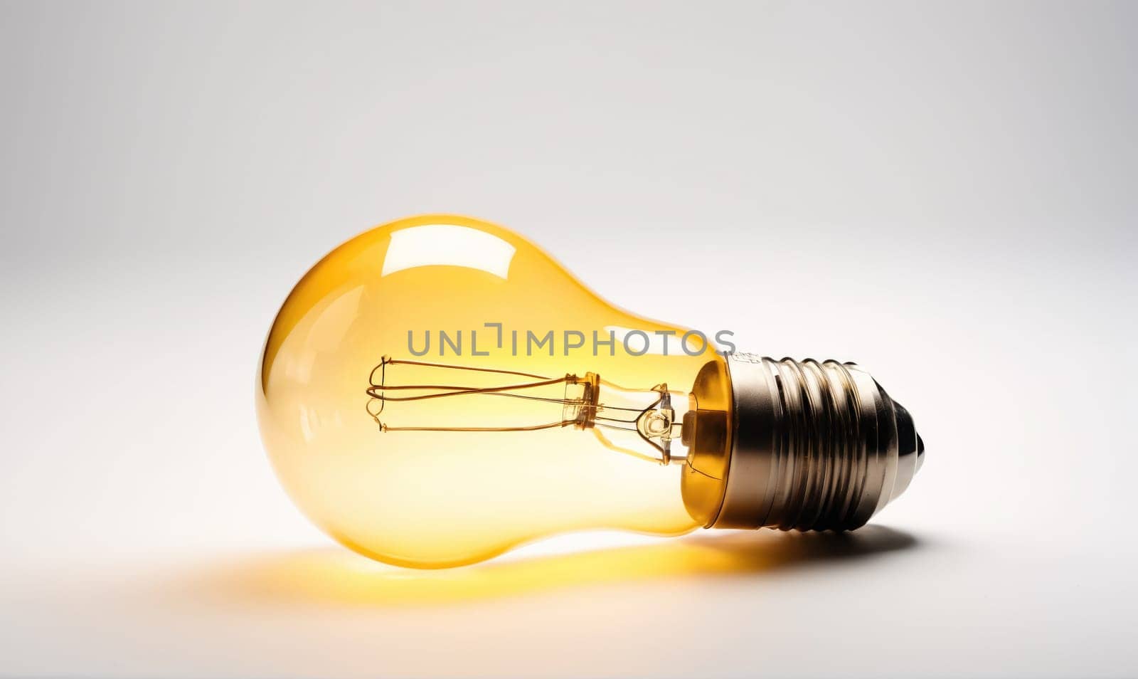 A light bulb lays on a white surface. The bulb is yellow and has a clear glass exterior, revealing the thin filament inside. The bulb is lying on its side, and the base is visible