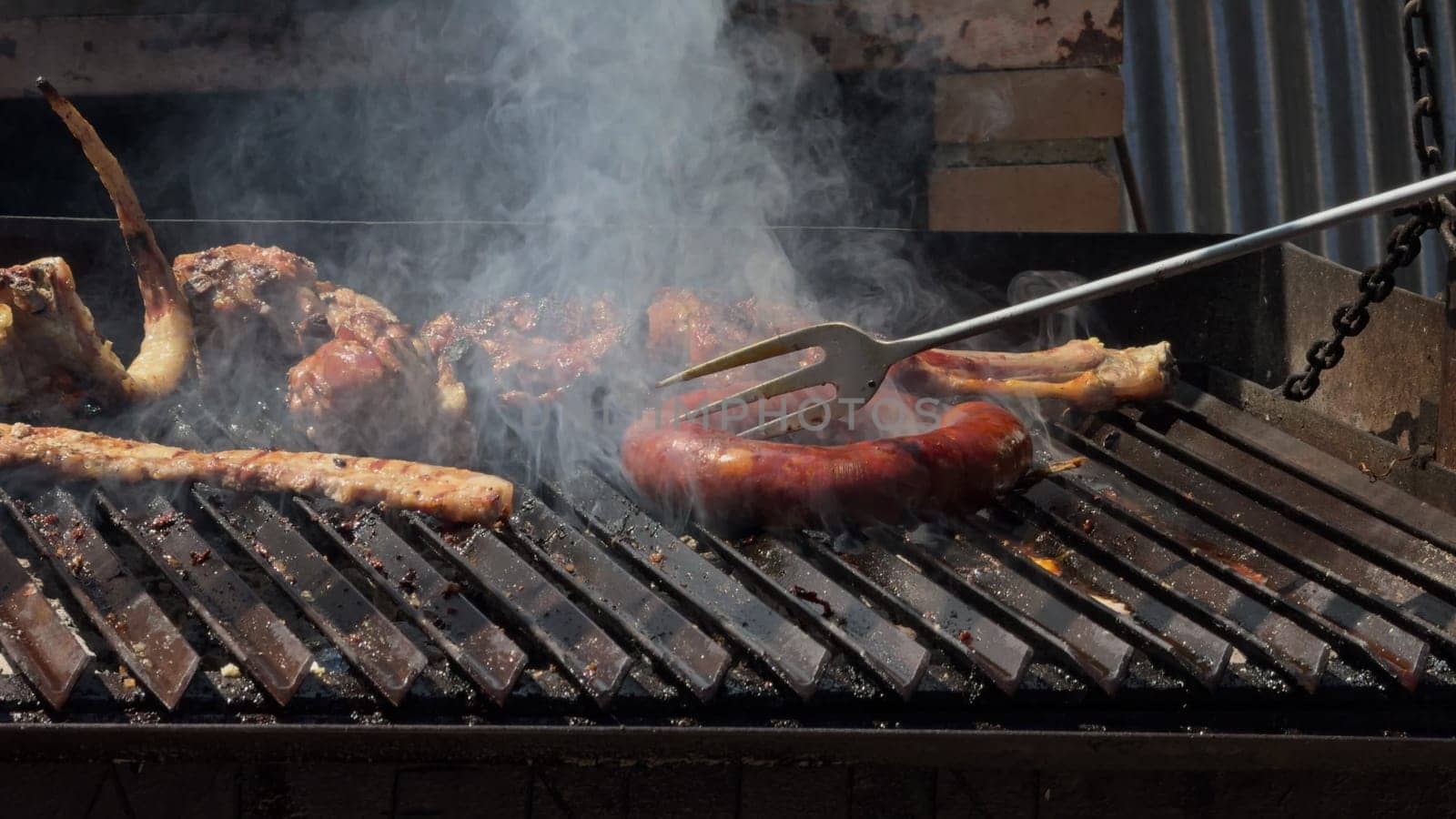 Video showing various meats being grilled on a barbecue, using a large fork to flip and check the doneness of the steaks over open flames and hot coals.
