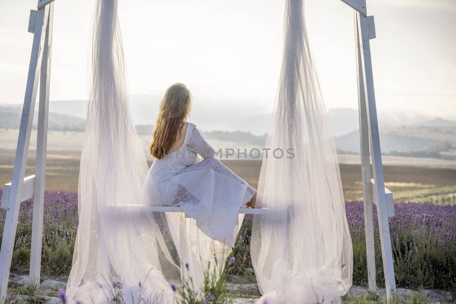 A woman is sitting on a bench under a white canopy. She is wearing a white dress and has long hair. The scene is set in a field with purple flowers