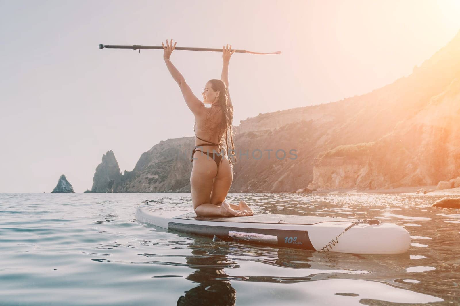A woman in a bikini kneels on a paddleboard, holding the paddle above her head, with a rocky coastline in the background.