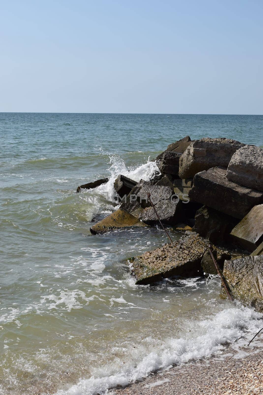 The waves of the turquoise sea break on the coastal boulders and a dense white foam is formed. Clear blue sky. White waves breaking on sandy beach through gap in rocks on sea