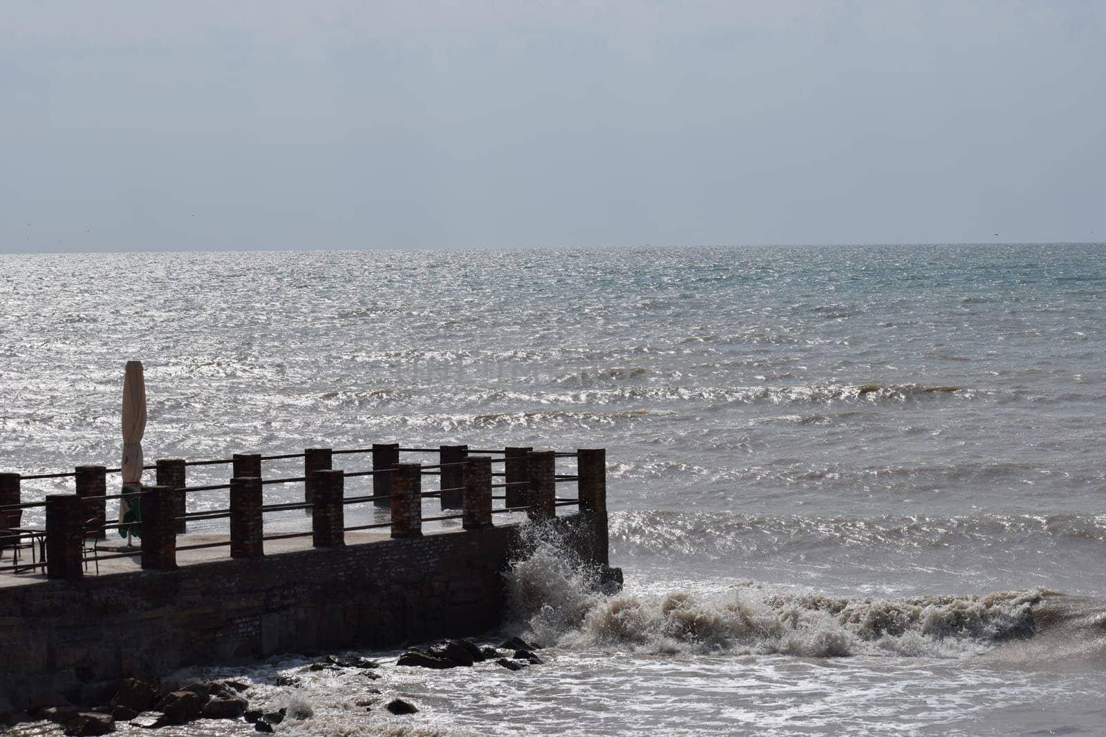big sea waves splashing against a pier. The pier cuts the sea waves off the coast during a stormy sunny day.