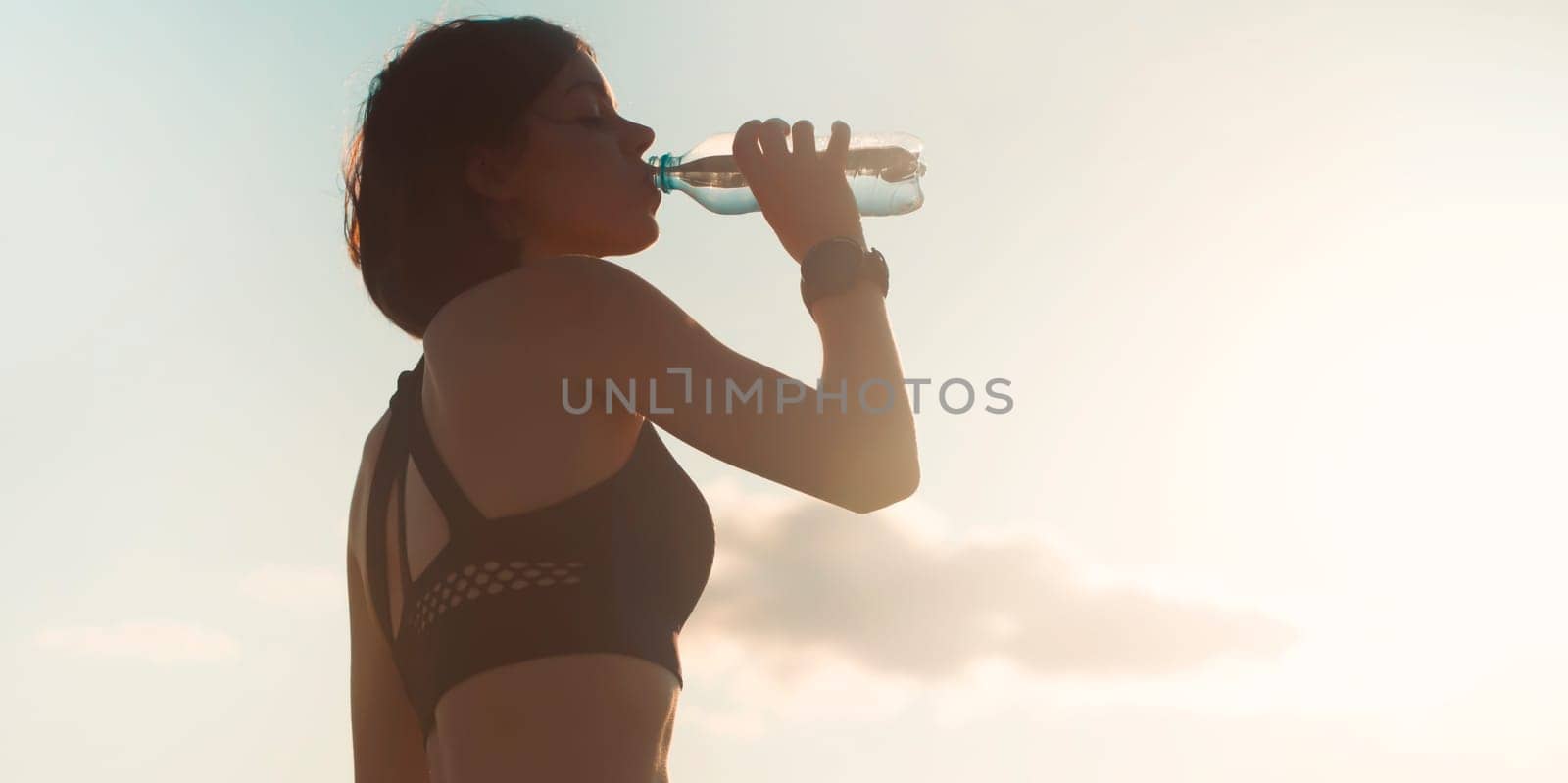 A young girl takes a break during an intense workout and jogging and drinks refreshing cool water from a bottle, a woman trains outdoor in the evening at sunset.