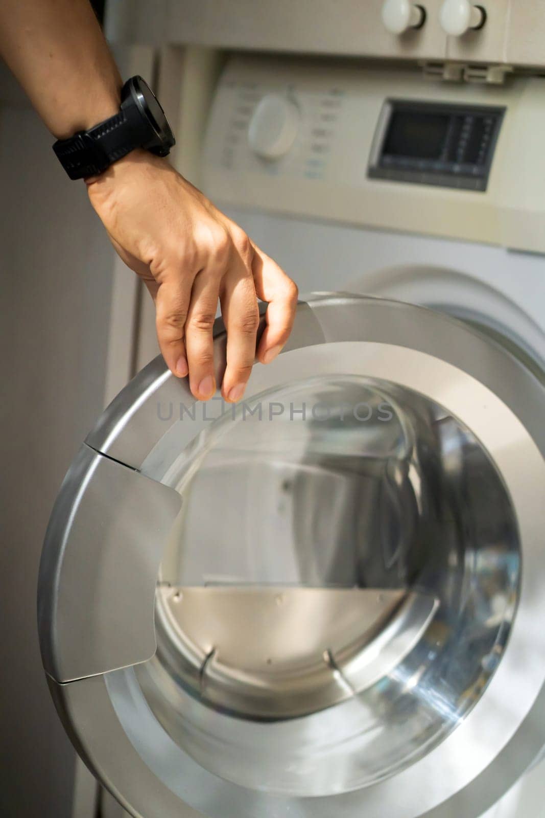 A male hand opens the door of a new washing machine in his modern, bright bathroom, close-up. The man washes clothes in the laundry.