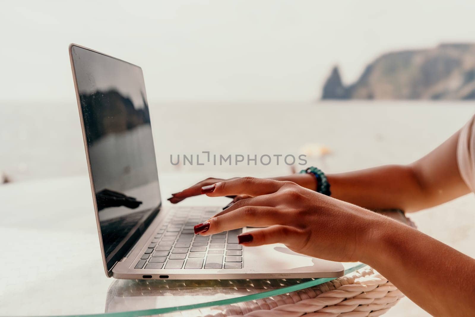 Close-up of hands typing on a laptop with a beach and ocean in the background.