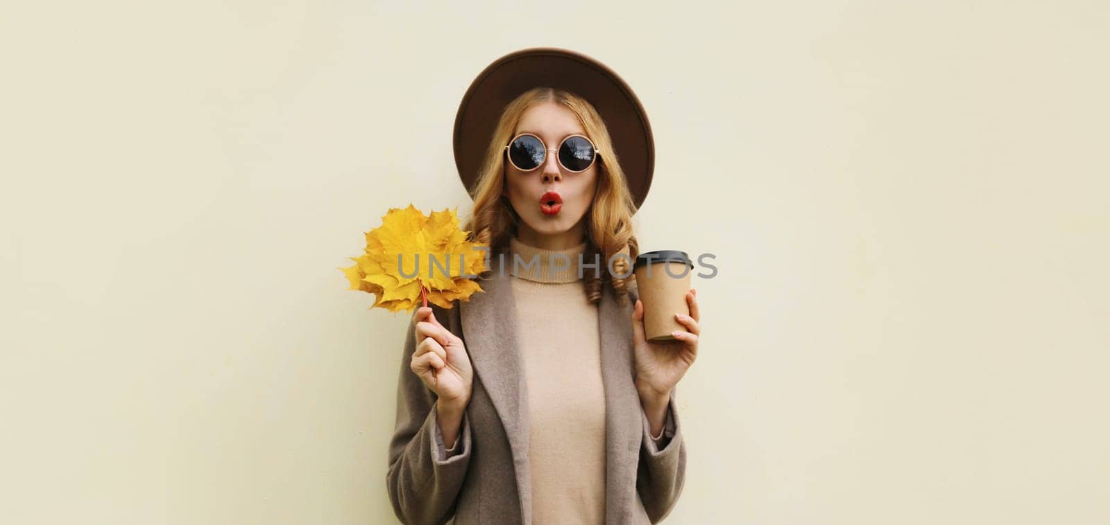 Autumn portrait of beautiful happy surprised woman with yellow maple leaves, stylish emotional girl holding cup of coffee posing isolated on brown background