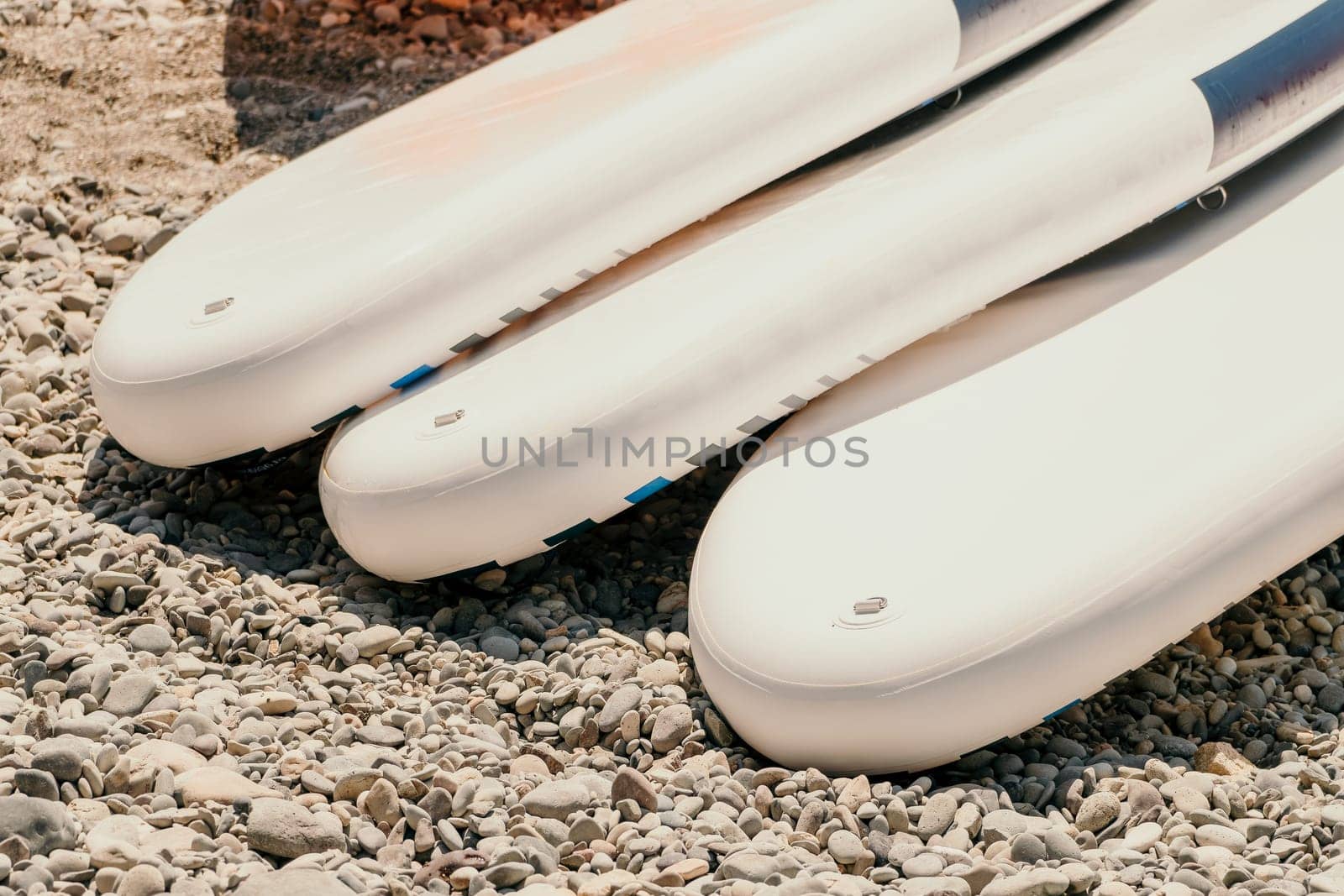 Three white inflatable paddle boards are stacked on a pebble beach. The boards are in focus, while the pebbles are out of focus.
