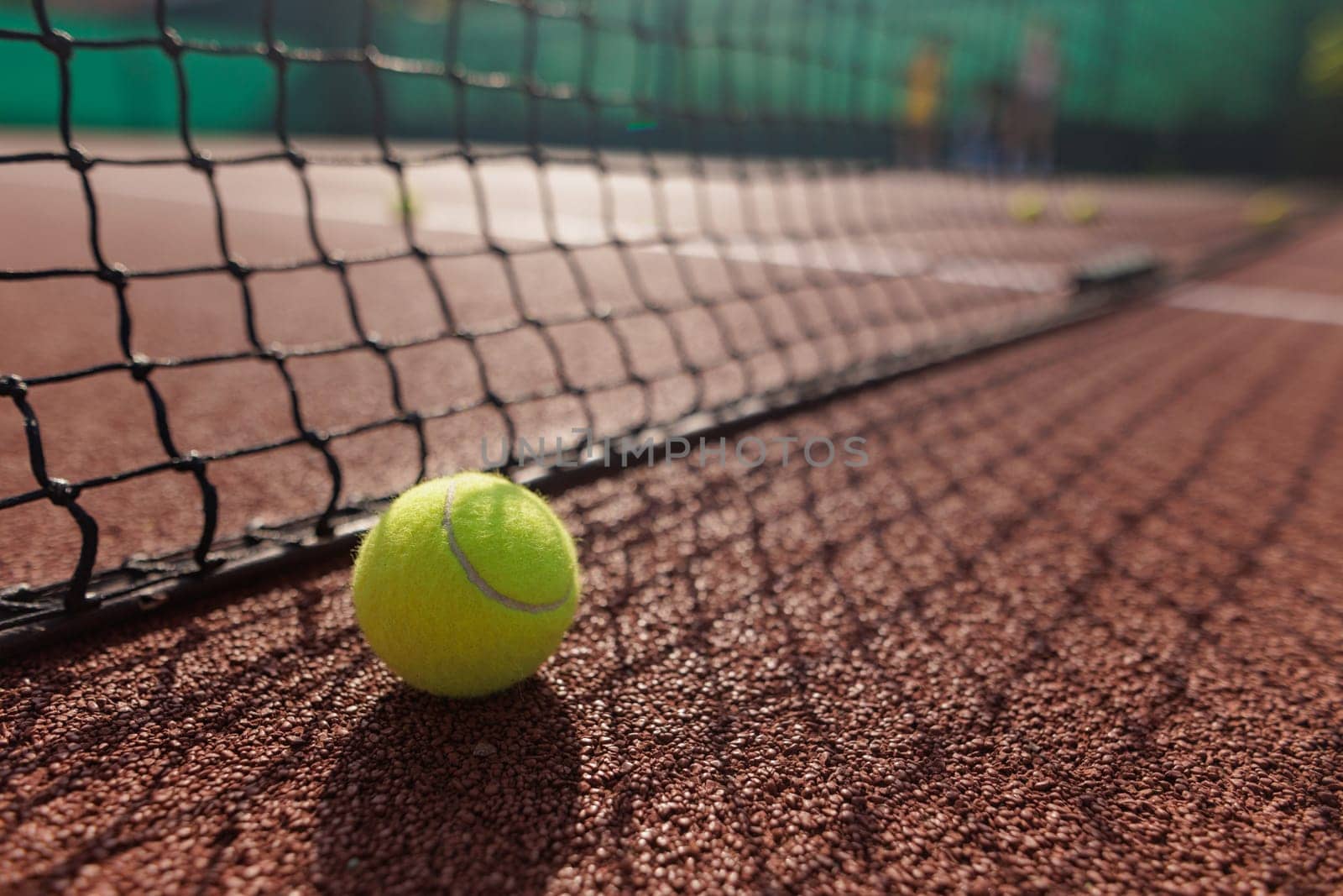 A close-up of a tennis ball resting on a clay court, with a net in the foreground and blurred players in the background