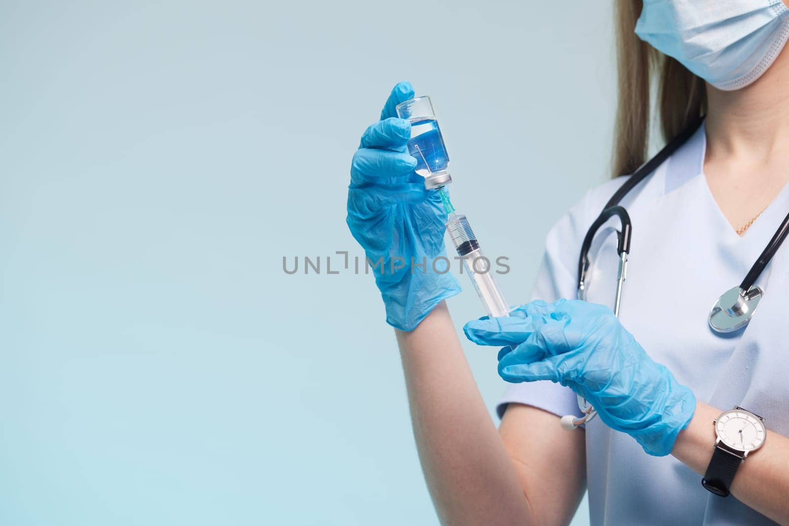 A healthcare professional in scrubs and gloves prepares a syringe with a blue liquid. They are wearing a mask and have a stethoscope around their neck, set against a light blue background.