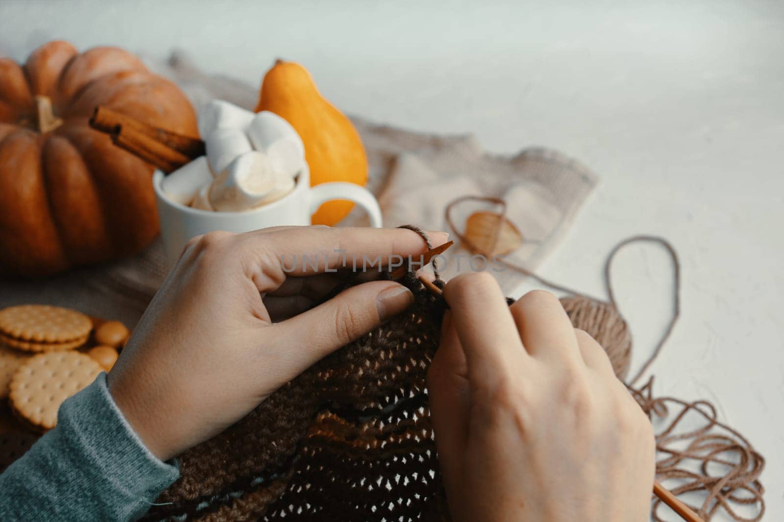 Hands are knitting among autumn decorations, biscuits, and a warm beverage, creating a cozy fall scene
