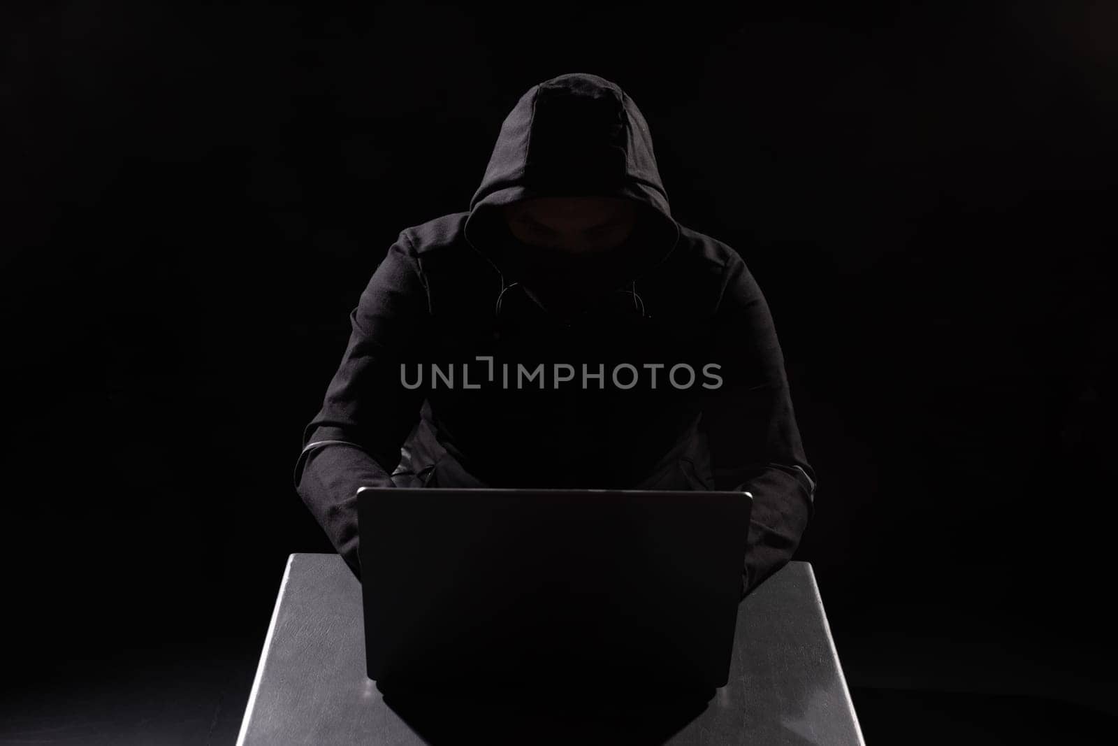 Cyber hacker in hood behind laptop in dark room, digital cyber security concept, computer hacking, information security