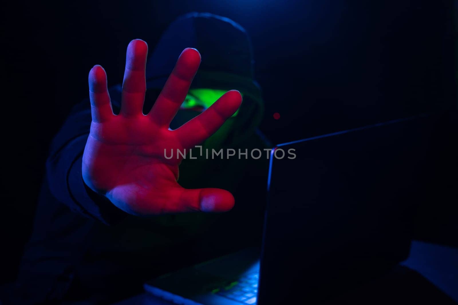 Cyber hacker in hood behind laptop puts hand forward hiding his face in dark room, concept of digital information protection, Computer hacking, stop cyber hacker