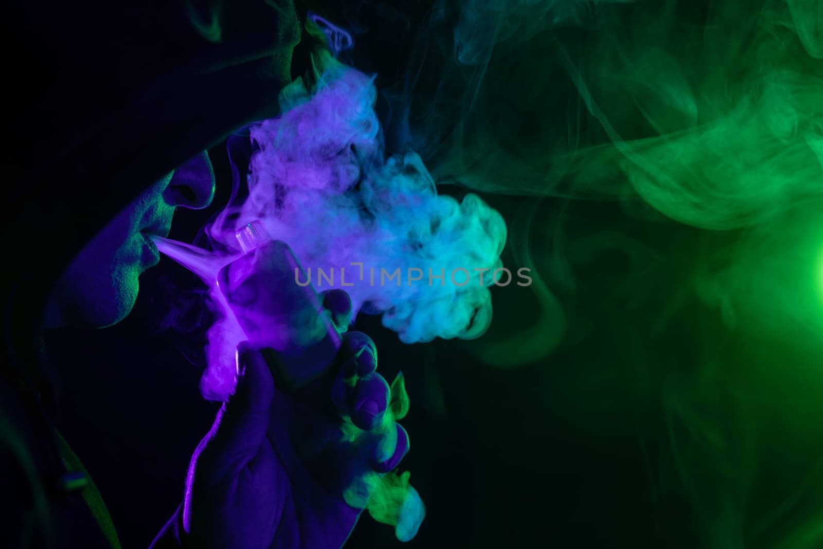 male vaper holding vape electronic cigarette close-up on background of colored smoke, concept of harm of smoking
