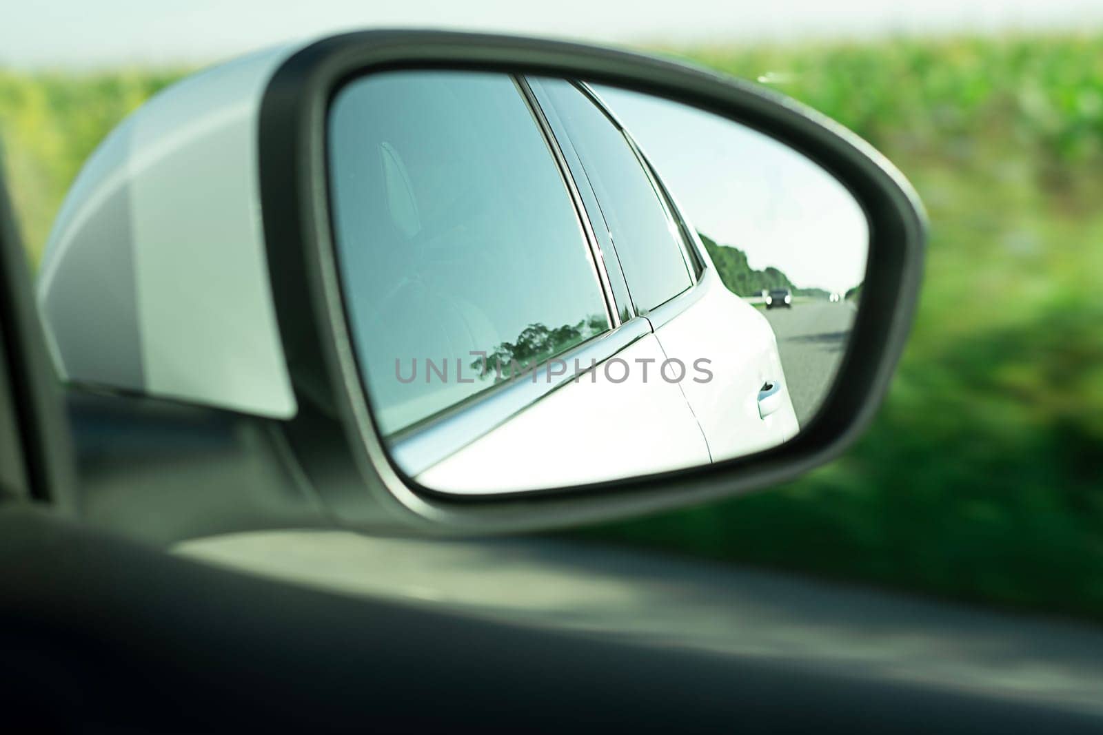 Rear-view mirror, close-up, with the reflection of a white car when the car is moving against the background of green trees. Soft focus. An essential element for driving safety.