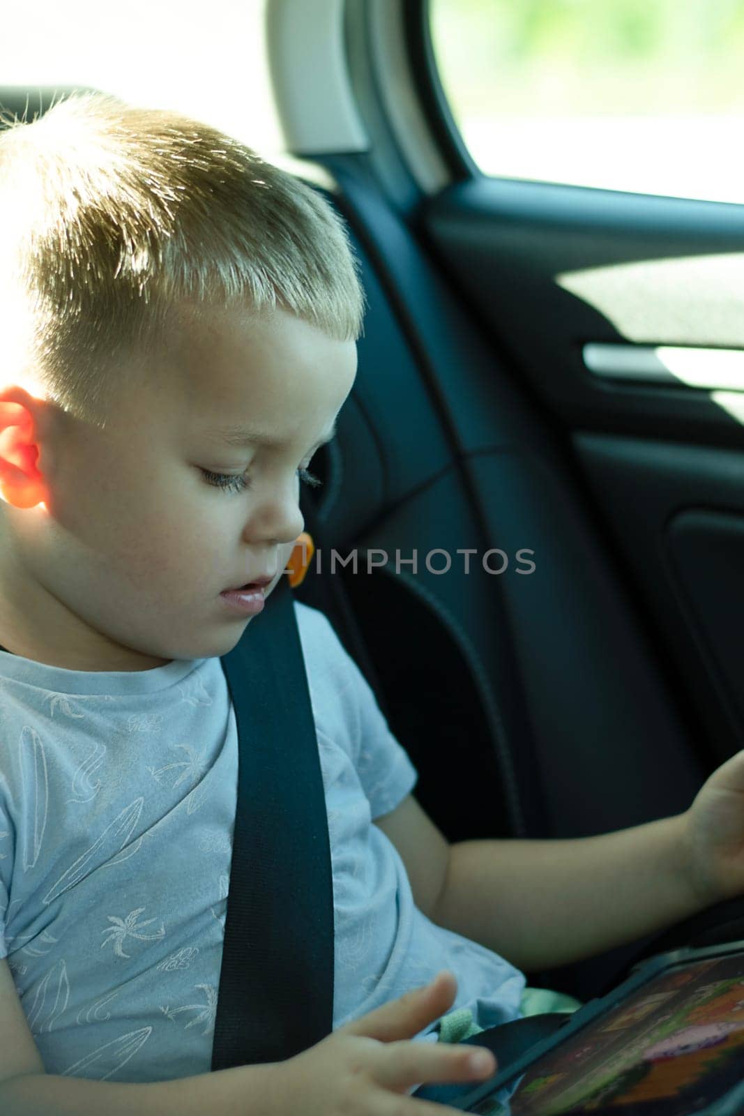 Security concept. A 4-year-old boy is riding in a car with a seat belt fastened in a child seat in the back seat. He travels with his family and watches cartoons on his iPad. Soft focus. vertical