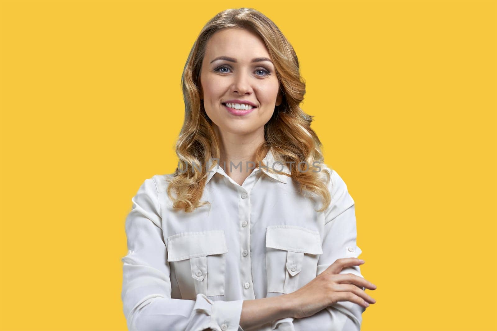 Protrait of a young blonde smiling woman with folded arms. Vivid yellow background.