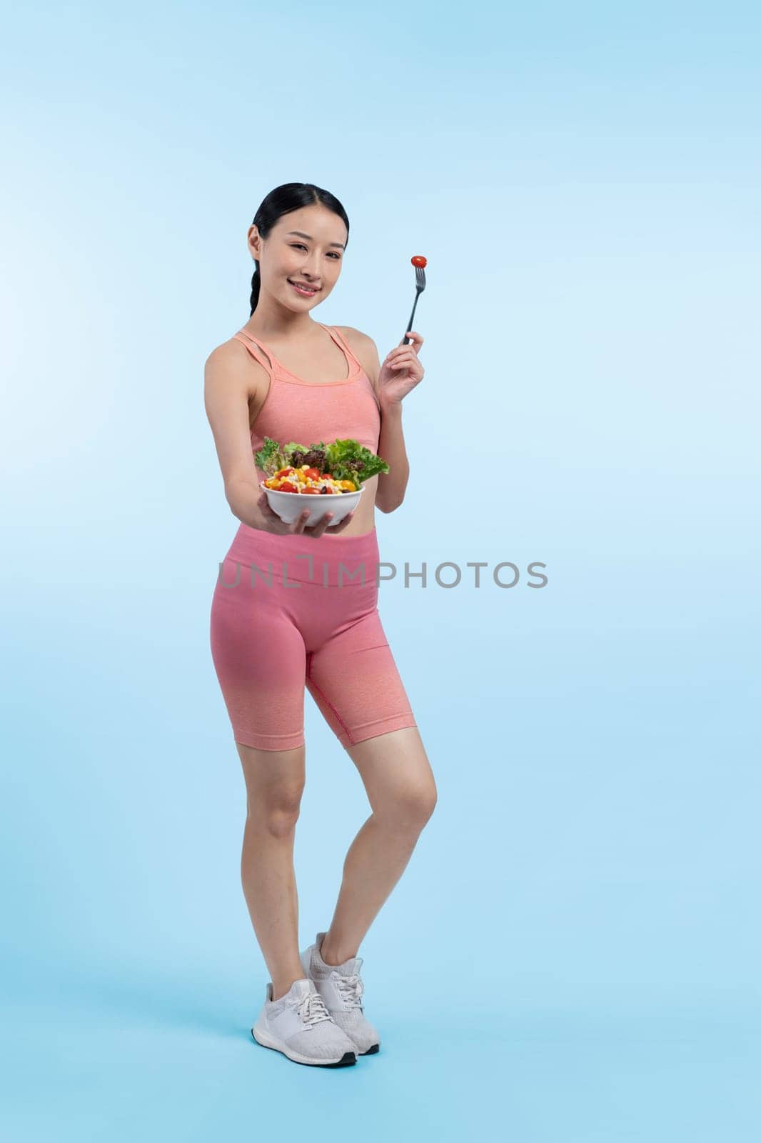 Young sporty Asian woman in sportswear holding salad bowl fill with vibrant of fruit and vegetable. Natural youthful and fit body lifestyle with balance nutrition on isolated background. Vigorous