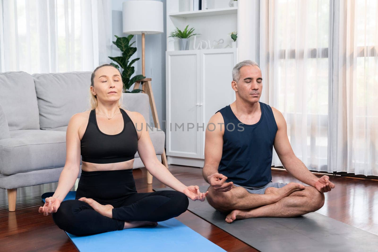 Happy active senior couple in sportswear being doing yoga in meditation posture on exercising mat at home. Healthy senior pensioner lifestyle with peaceful mind and serenity. Clout