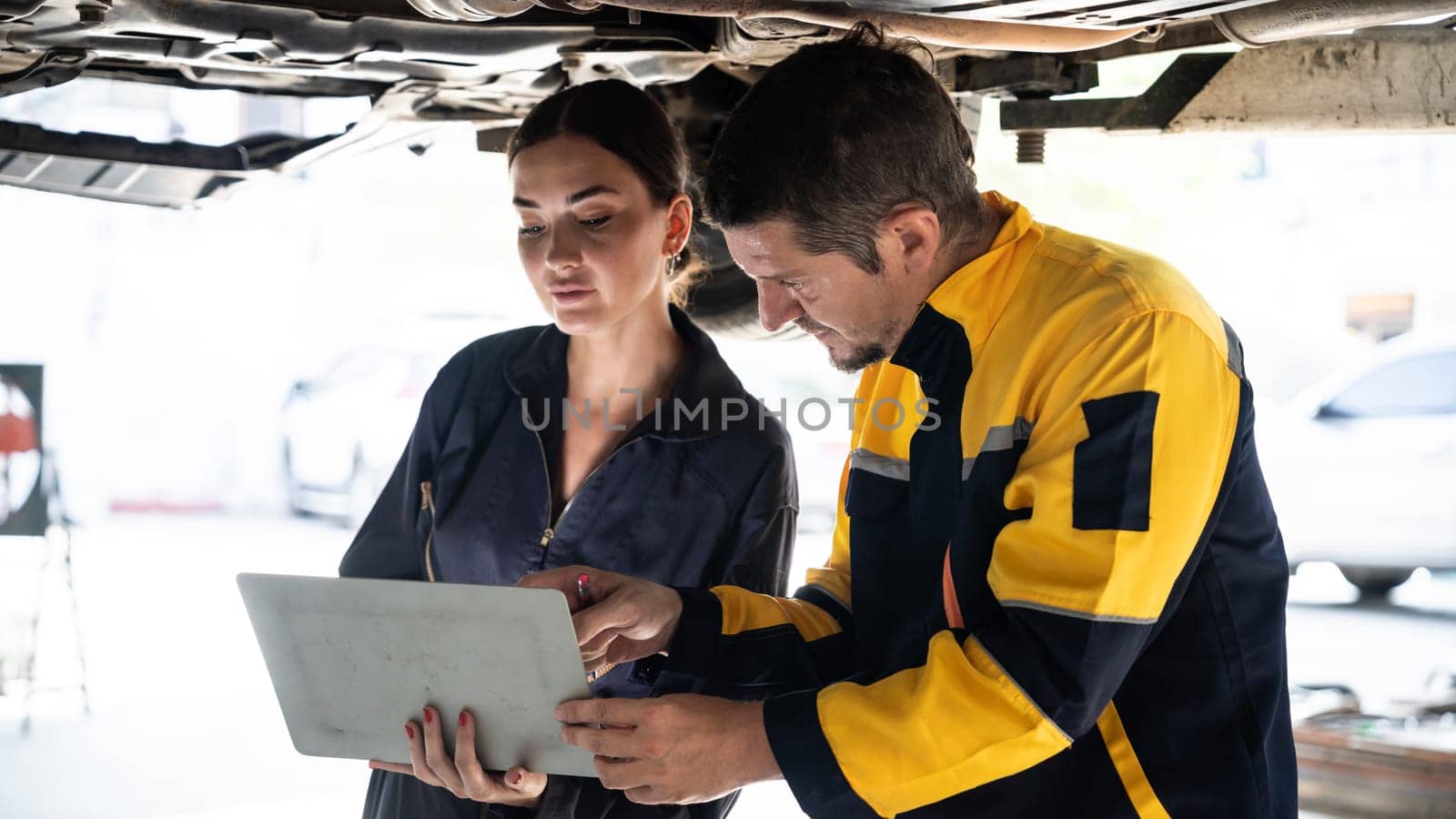 Two vehicle mechanic working together underneath lifted car, conduct car inspection with laptop. Automotive service technician in uniform carefully make diagnostic troubleshooting. Panorama Oxus