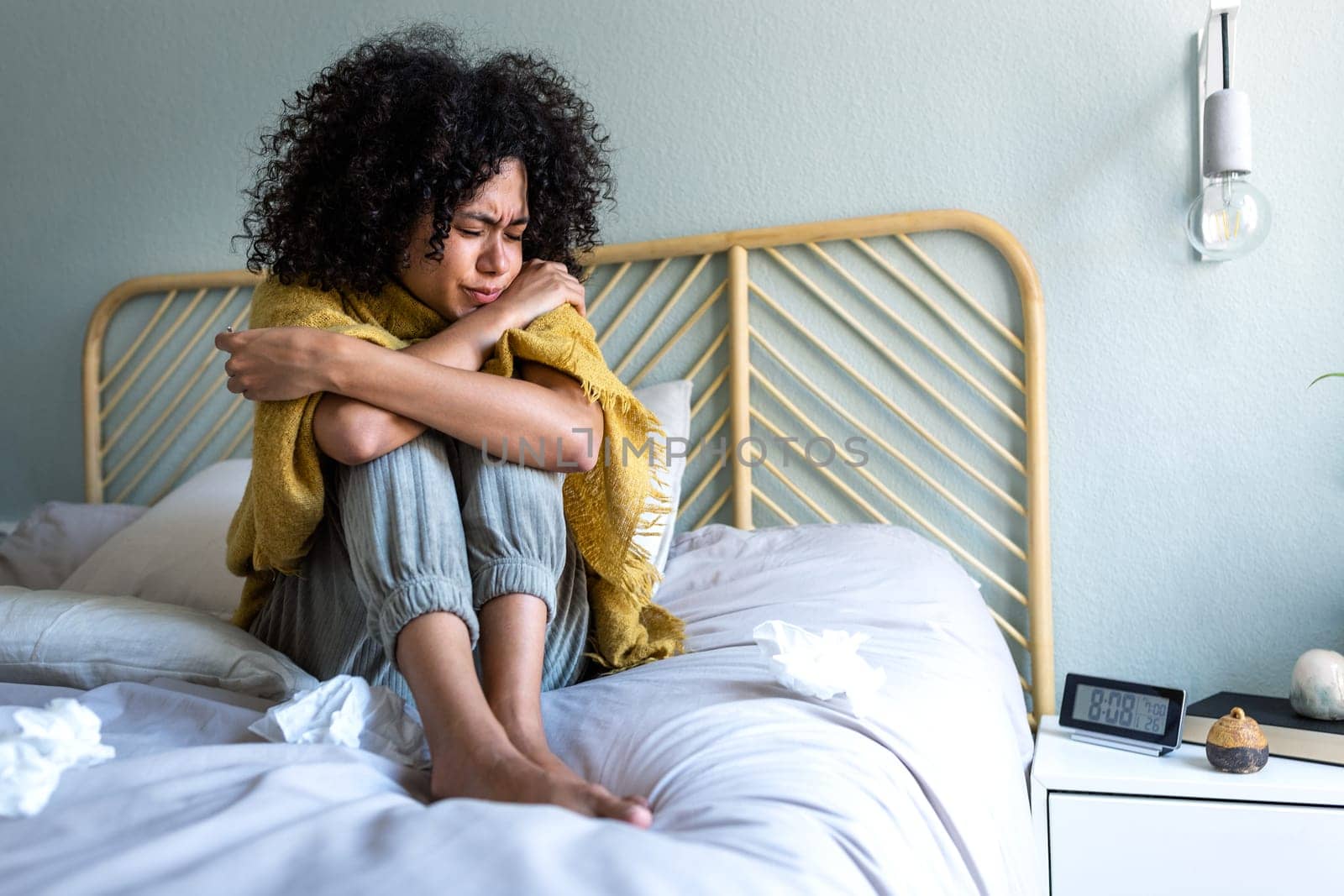 Young multiracial woman feeling sick and cold sitting on bed covered with blanket, choking temperature with thermometer. by Hoverstock