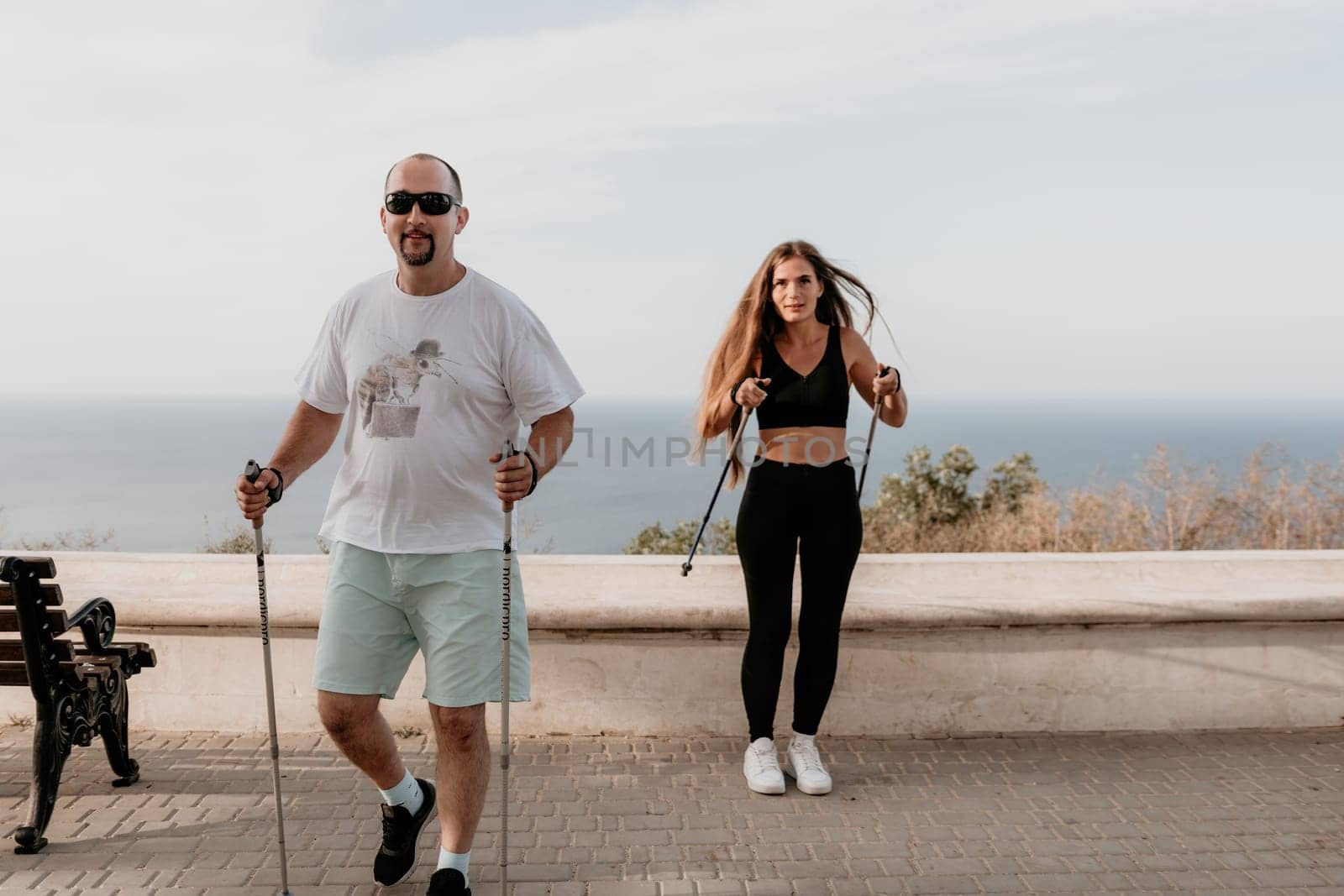 Happy Middle aged couple or friends practicing nordic walking in park near sea. Mature couple with trekking poles walking, practicing Nordic walking outdoors. Aging youthfully and sport concept.