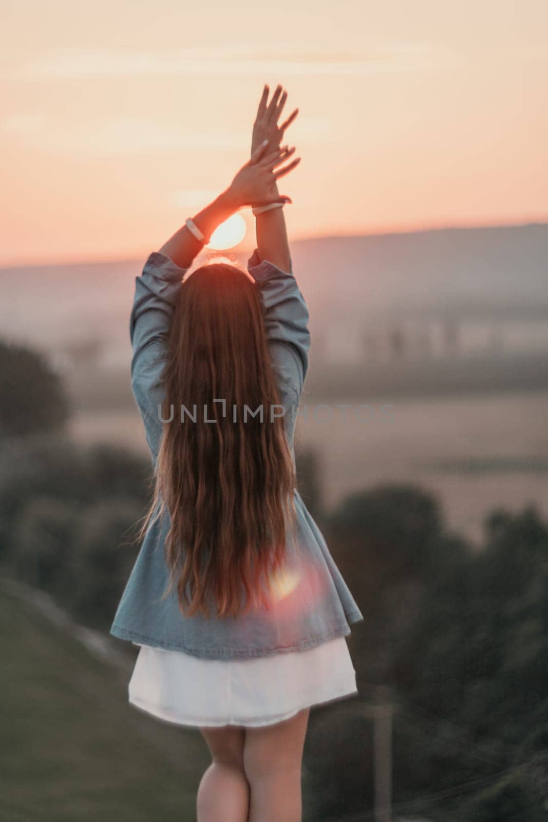 Happy woman standing with her back on the sunset in nature in summer with open hands. Romantic beautiful bride in white boho dress posing with mountains on sunset by panophotograph