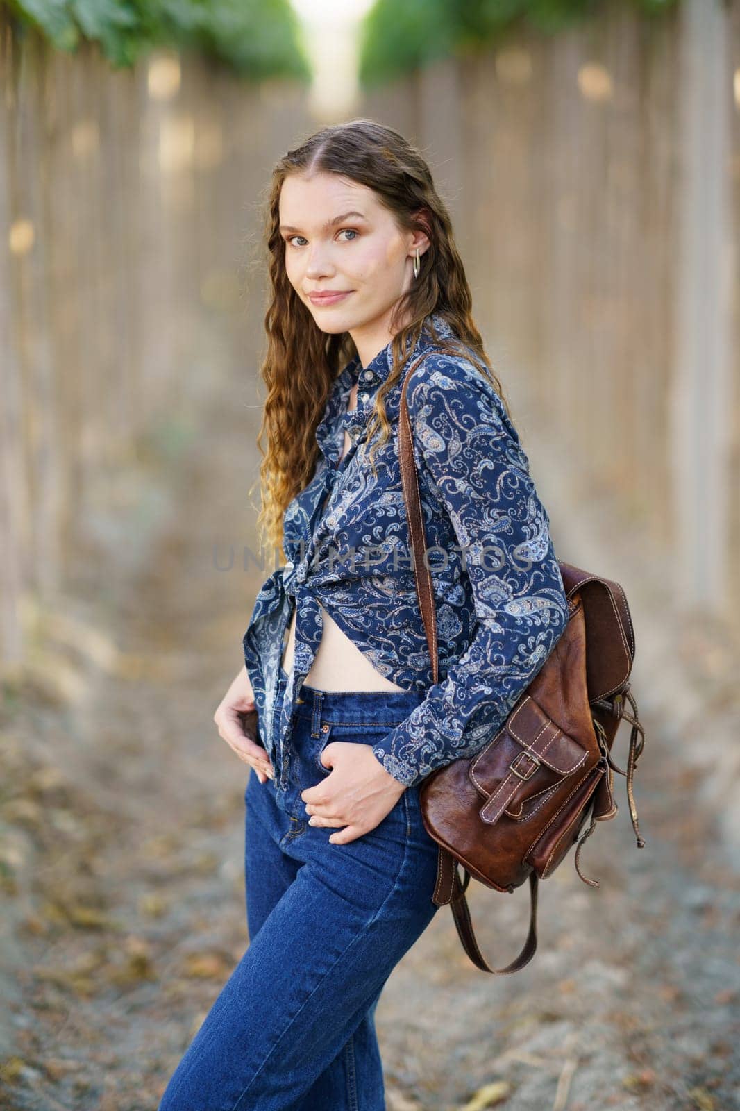 A young woman stands confidently in a beautiful vineyard, dressed in a stylish blue floral shirt paired with denim pants, while carrying a chic brown backpack that complements her outfit
