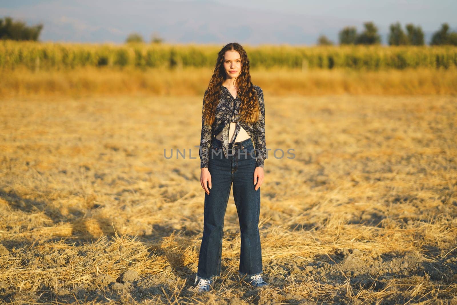 A young woman with long, curly hair stands proudly in a dry, golden field. The serene landscape features distant trees under a clear blue sky, creating a breathtaking and inviting scene