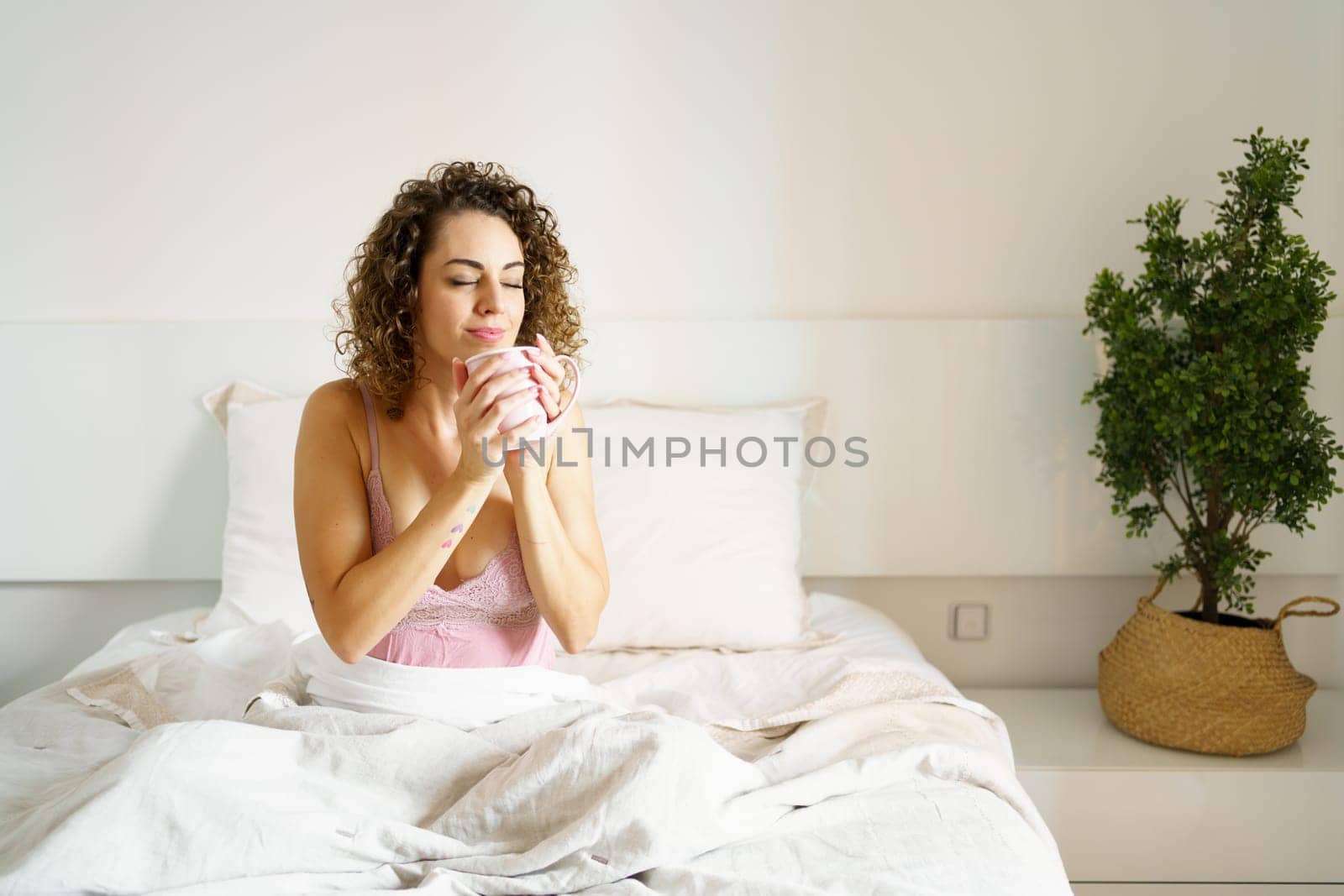 Relaxed young woman in nightwear holding fresh aromatic coffee mug while sitting in bed at home