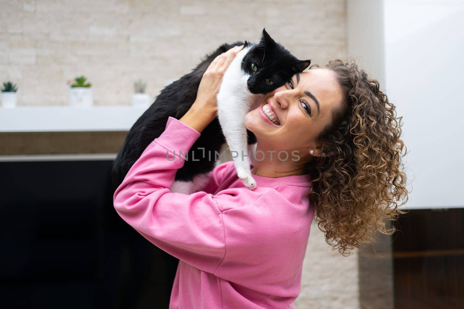 Side view of young female with curly hair in pink casual cloth, hugging adorable cat with black and white fur and looking at camera while in modern apartment
