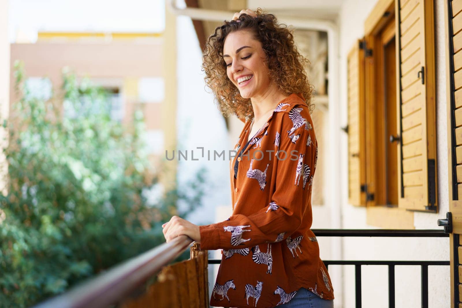 Side view of young female in stylish outfit smiling and looking down while standing on balcony against blurred background